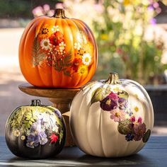 three painted pumpkins sitting on top of a wooden table in front of some flowers