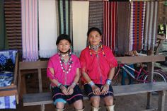 two women sitting next to each other on a bench in front of some colorful fabrics