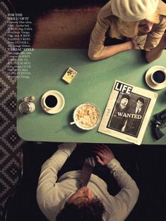 two people sitting at a green table with coffee and magazines on it, one person has his head down