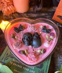 two skulls in a heart shaped bowl filled with pink liquid and flowers, surrounded by candles