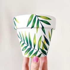 a woman's hand holding up a white cup with green leaves painted on it