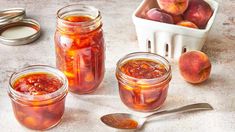 three jars filled with peach jam next to two spoons and a container full of peaches