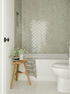 a white toilet sitting next to a bath tub under a window in a bathroom with gray tiles on the walls