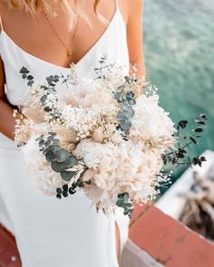 a woman in white dress holding a bouquet of flowers and greenery next to water