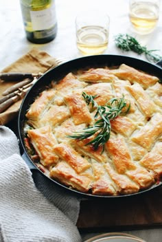 a pie in a pan with rosemary garnish on top and wine glasses next to it