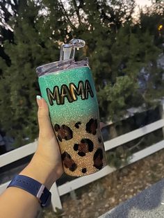 a person holding up a cup with the word mama on it in front of some trees