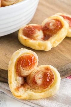 two heart shaped pastries sitting on top of a wooden cutting board next to a white bowl