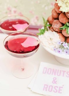 two glasses filled with red liquid next to donuts and roses on a white table