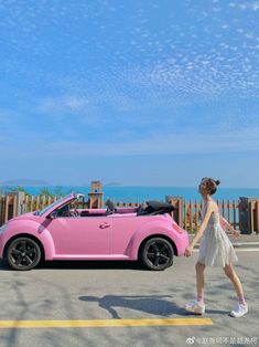 a woman walking next to a pink car on the road near the ocean and fence