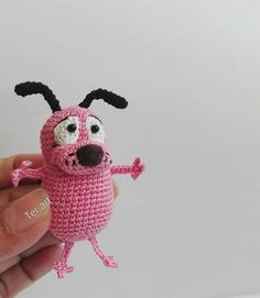 a crocheted pink toy with black ears and legs is being held up by someone's hand