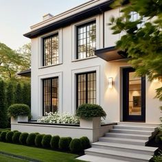 a white house with black windows and steps leading up to the front door is surrounded by greenery