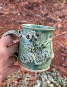 a hand holding a green ceramic mug with an animal on it's face and eyes