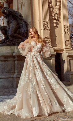 a woman standing in front of a statue wearing a wedding dress with long sleeves and flowers on it