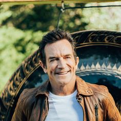 a man in a brown leather jacket smiles at the camera while standing next to a tire