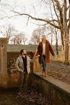 a man and woman holding hands while standing on a stone wall in front of trees