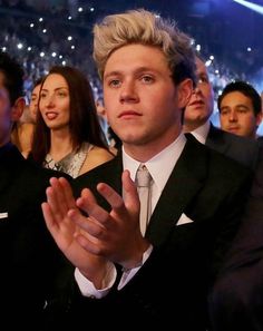 a man in a suit and tie clapping with other people watching from the stands behind him