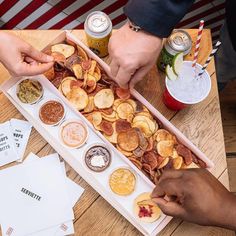 two people are grabbing chips from a tray on a table with other snacks and condiments