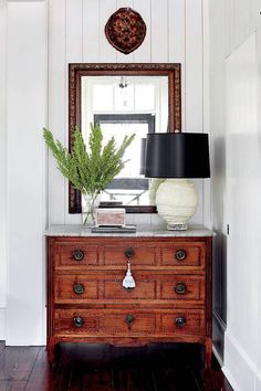 a wooden dresser with a lamp on top of it next to a mirror and plant