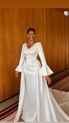 a woman in a white dress standing next to a wooden wall with her hands on her hips