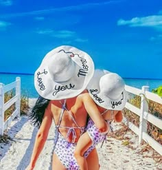 two women in hats walking on the beach with their arms around each other's shoulders