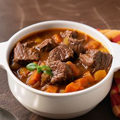 a white bowl filled with beef and carrots on top of a wooden table next to a spoon