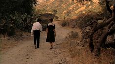 two people walking down a dirt road in front of some trees and hills with mountains in the background