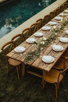 a long wooden table with white plates and flowers on it in front of a pool