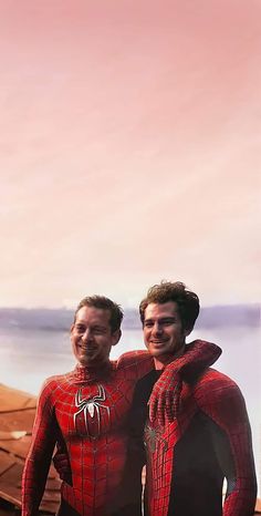 two men in spider - man suits standing next to each other near the ocean, smiling