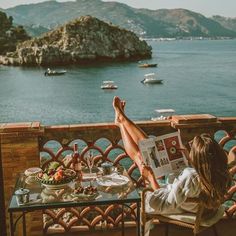 a woman sitting at a table reading a magazine overlooking the ocean and boats in the water