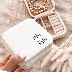 a person holding an open white box with jewelry inside it on top of a bed