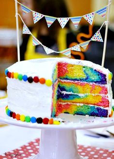 a rainbow cake with white frosting and multi colored icing on a table next to a bunting banner