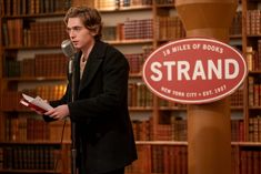 a man standing at a microphone in front of a book shelf with books on it