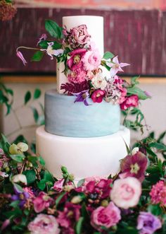 a white and blue wedding cake with flowers on the bottom tier, surrounded by greenery