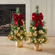 two small christmas trees with red bows and gold ornaments are sitting on the floor in front of a fireplace