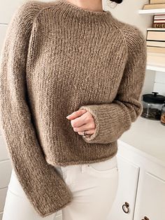 a woman wearing a brown sweater and white pants standing in front of a book shelf