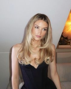 a woman with long blonde hair sitting on a couch in a black dress and looking at the camera