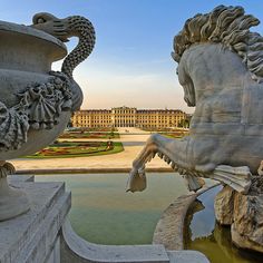 two statues in front of a large building with a fountain next to it and an ornate garden behind them