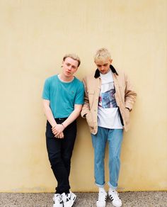 two young men standing next to each other in front of a yellow wall with their hands on their hips