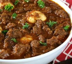 a white bowl filled with meat and potatoes on top of a red and white table cloth