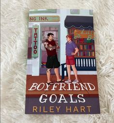 the book boyfriend and girlfriend goals by riley hart is laying on a white carpet with an image of two men standing in front of a tattoo parlor