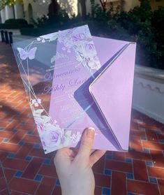 a person holding up two purple and white wedding cards in front of a brick walkway