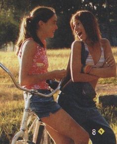 two young women sitting on top of a bike talking to each other in the grass