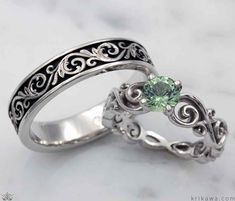 two wedding rings with green stone in the center on a white marble surface, one is made from silver and the other has an intricate fili