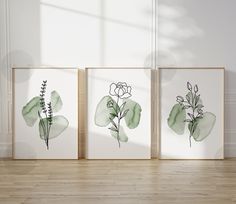 three watercolor paintings on white paper with green leaves and flowers in the middle, against a light wood floor