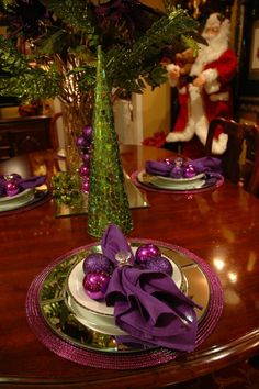 the table is set for christmas dinner with purple ornaments and greenery in a vase