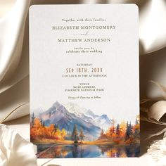 a wedding card with an image of mountains and trees on it, in front of white satin