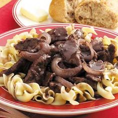 a red plate topped with pasta and meat covered in sauce next to a loaf of bread