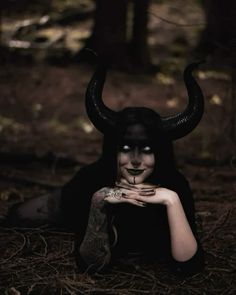 a woman with black makeup and horns on her face is laying in the woods while holding her hands to her chest