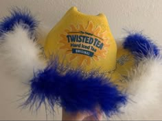 a person holding up a yellow and blue hat with white feathers on the bottom, which reads twisted tea hard boiled tea