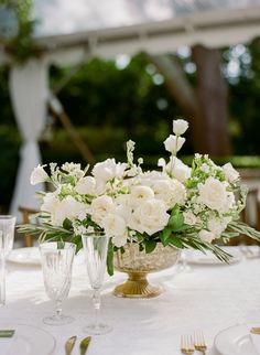 a table with white flowers and greenery in a gold vase on top of it